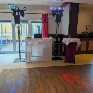 Professionelles und sauberes DJ-Setup von PvD - Events & Technik im Festsaal des Hotels Weserschiffchen in Porta Westfalica, ausgestattet mit LED-Lichtstativen, Moving Heads und Kompakt-Lautsprechern direkt vor den Türen zur Außenterrasse.