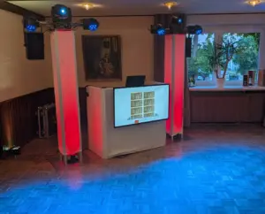 Elegantes weißes DJ-Pult von PvD - Events & Technik bei einer Hochzeit in Bohmte mit integriertem TV-Bildschirm für die Fotobox-Diashow und rot beleuchteten Säulen mit Moving Heads.