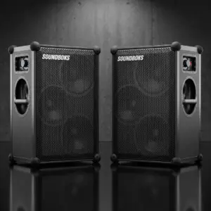 Zwei kabellose SOUNDBOKS 4 Akku-Bluetooth-Lautsprecher im Stereo-Set nebeneinander platziert, mit schwarzem IP65-Gehäuse, Frontgitter und Lautstärkeregler vor edlem Studiolook-Hintergrund.