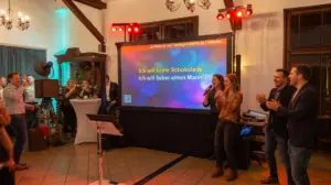 Gut gelaunte Gäste singen und feiern ausgelassen bei einem Event im Kreis Herford mit dem professionellen Karaoke-Miet-Setup von PvD - Events & Technik. Zu sehen ist eine 240 cm breite Rückprojektions-Leinwand mit Songtext, dazu Funkmikrofone, Säulen-PA, Steuerungspult und eine Fotobox in einem rustikalen Festsaal.